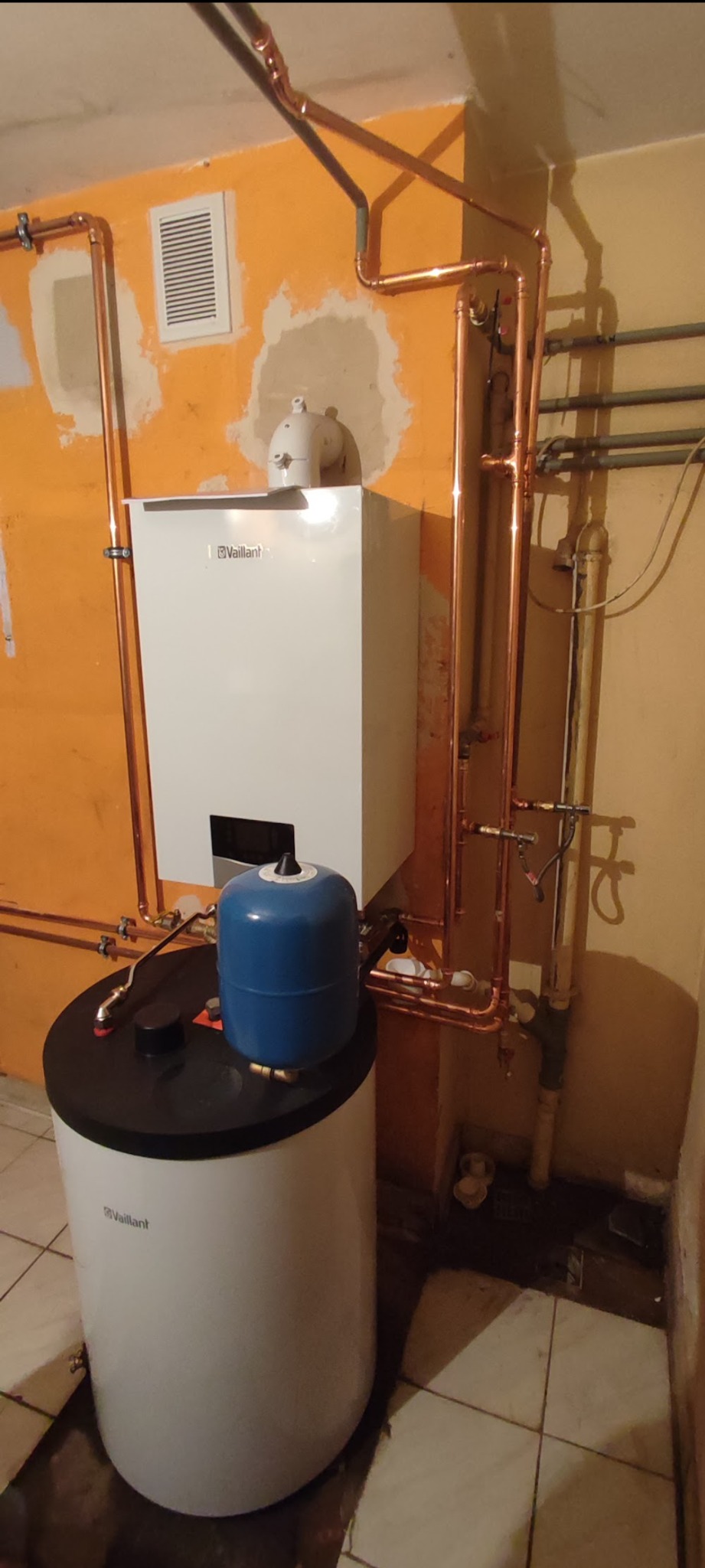 Biały piec gazowy Vaillant z miedzianą instalacją na pomarańczowej ścianie z niedokończonym wykończeniem. Obok biały bojler z niebieskim naczyniem wzbiorczym.