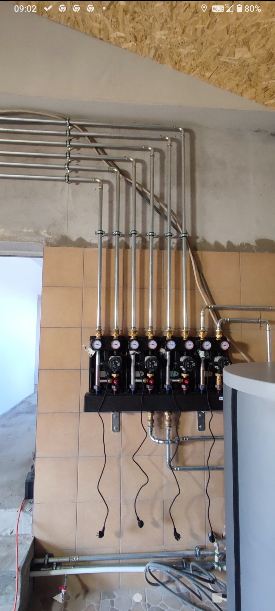 Zamontowana instalacja grzewcza z rzędami rur i pomp obiegowych na brązowych płytkach. Widoczne przewody elektryczne i fragment zbiornika. Surowy stan wykończenia.