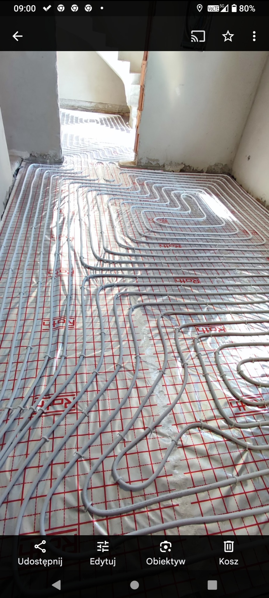 Ułożona instalacja ogrzewania podłogowego na folii z czerwoną kratką, widok z góry, w tle schody w surowym stanie, jasne światło.