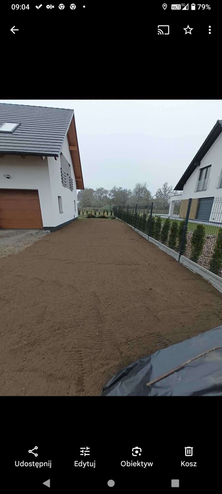 Przygotowany, wyrównany teren pod trawnik między dwoma domami jednorodzinnymi, ogrodzony siatką i tujami. Widoczne ślady po pracy glebogryzarki.