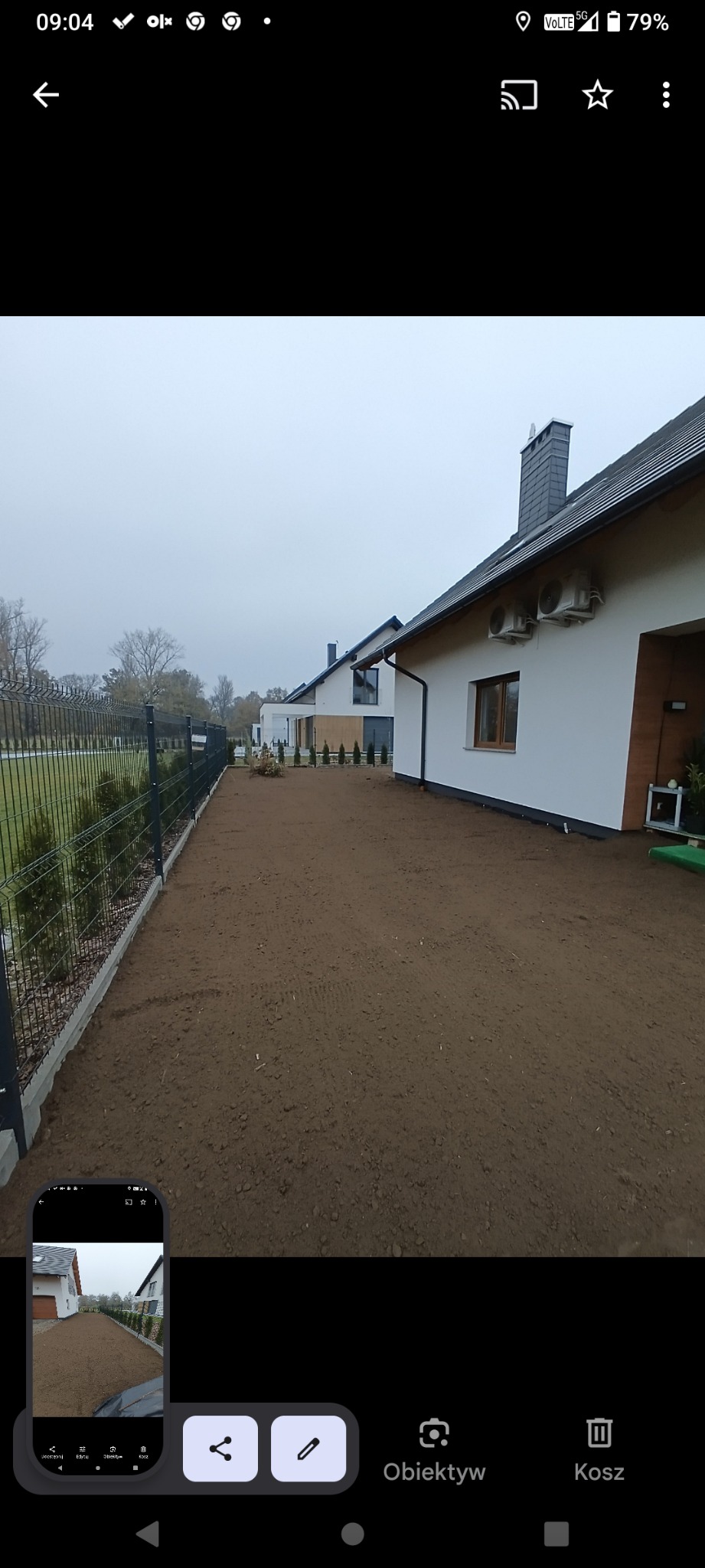 Przygotowany teren pod trawnik przy nowoczesnych domach jednorodzinnych, widok z perspektywy gruntu. Ogrodzenie z siatki, małe drzewka, pochmurne niebo.