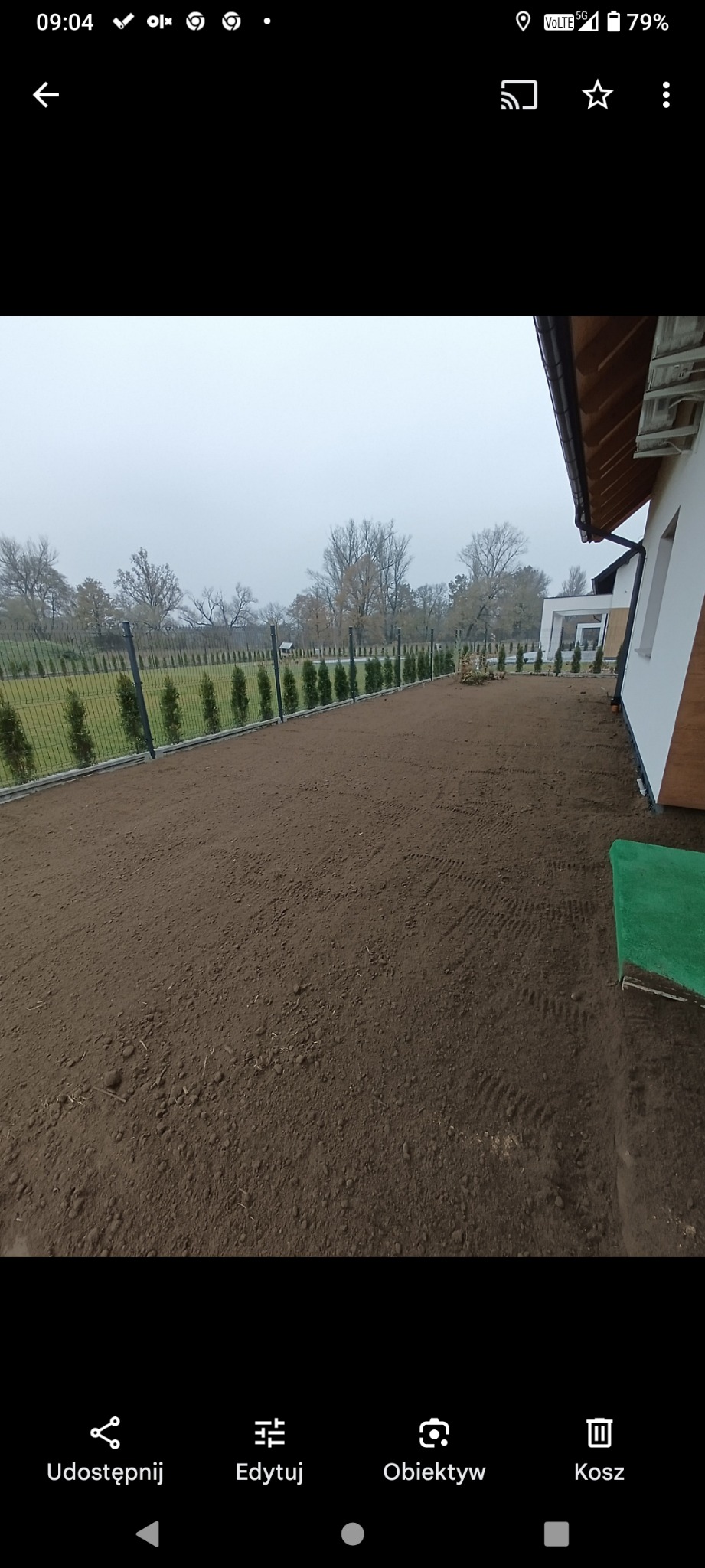 Niwelacja terenu: wyrównana ziemia pod trawnik, widoczne ślady opon, ogrodzenie z tujami, fragment budynku z drewnianym wykończeniem elewacji w tle.