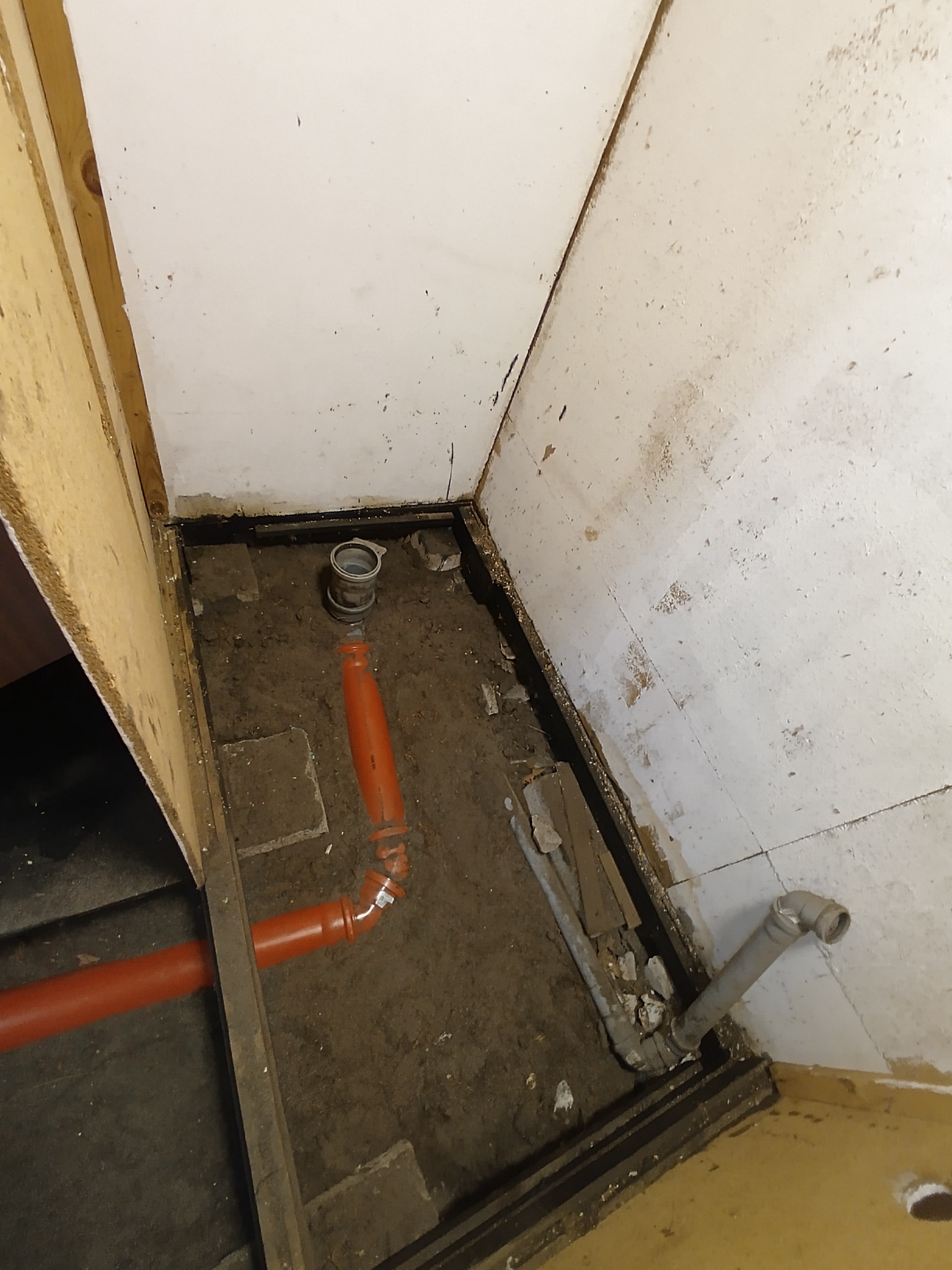 Fragment podłogi z instalacją kanalizacyjną w trakcie montażu. Widoczne rury PCV w kolorze pomarańczowym i szarym. Podłoże ziemne, ściany w stanie surowym.