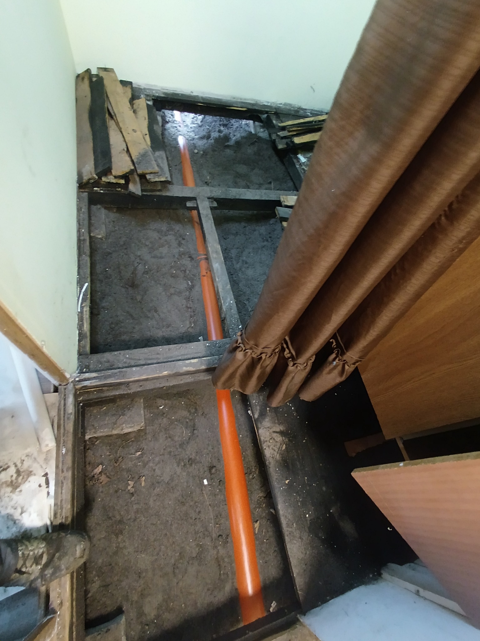 Pomarańczowa rura kanalizacyjna ułożona w wykopie pod podłogą, widoczne deski i fragment zasłony. Prace instalacyjne w trakcie remontu.