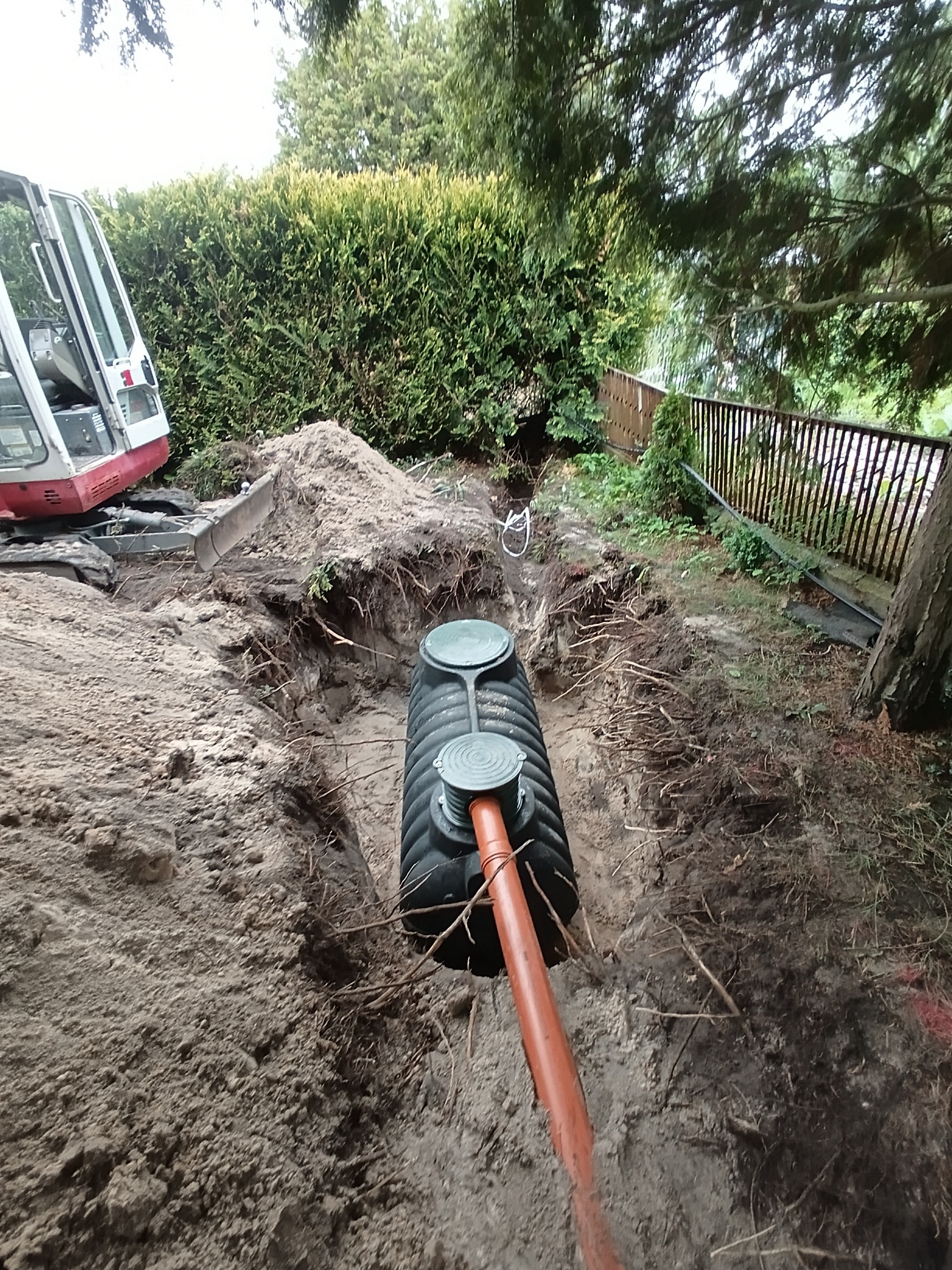 Montaż szamba betonowego w wykopie, widoczny fragment rury odprowadzającej i koparka w tle. Teren zielony z żywopłotem.
