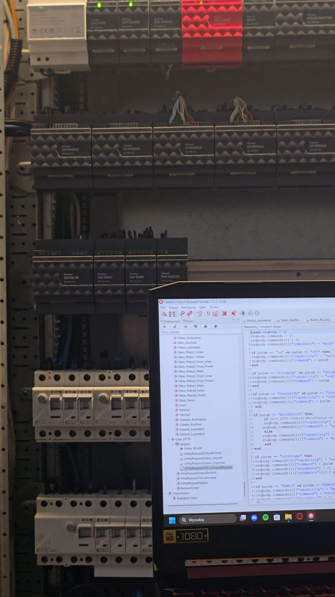 Panel sterowania z modułami Greton, widoczne kable i zabezpieczenia. Ekran laptopa z interfejsem programowania. Automatyka budynkowa w trakcie konfiguracji.