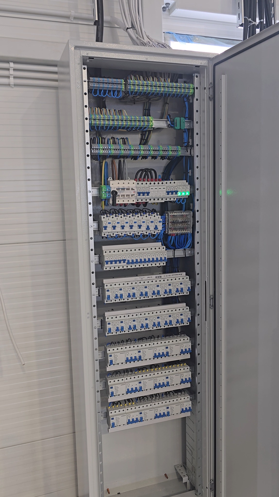 Otwarta szafa sterownicza z rozbudowaną instalacją elektryczną, zawierająca liczne zabezpieczenia i złącza, w pomieszczeniu o jasnych ścianach.