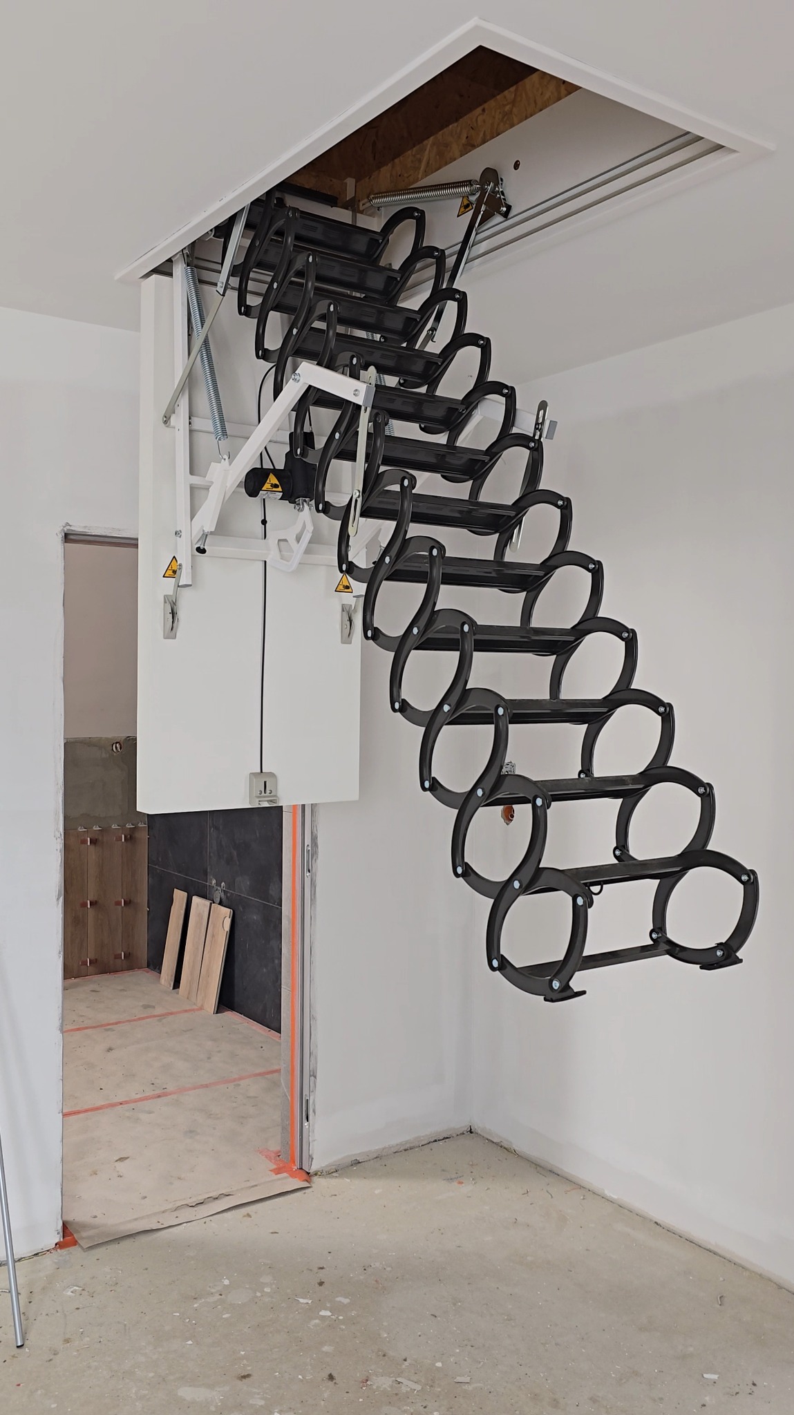Rozkładane, czarne schody strychowe z białym mechanizmem i klapą, zamontowane w suficie, widoczne w surowym pomieszczeniu w trakcie remontu.