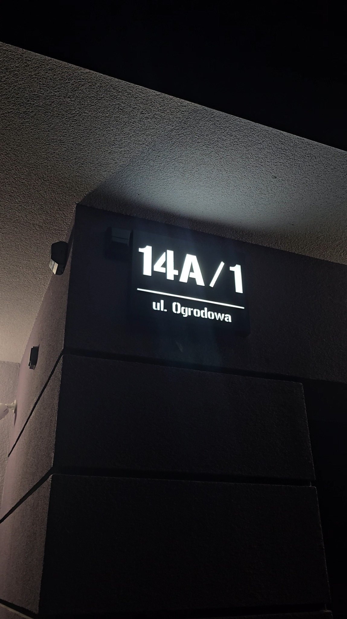 Podświetlany numer budynku 14A/1 z nazwą ulicy 'ul. Ogrodowa' w nowoczesnym stylu, zamontowany na elewacji budynku. Nocne ujęcie z dołu.