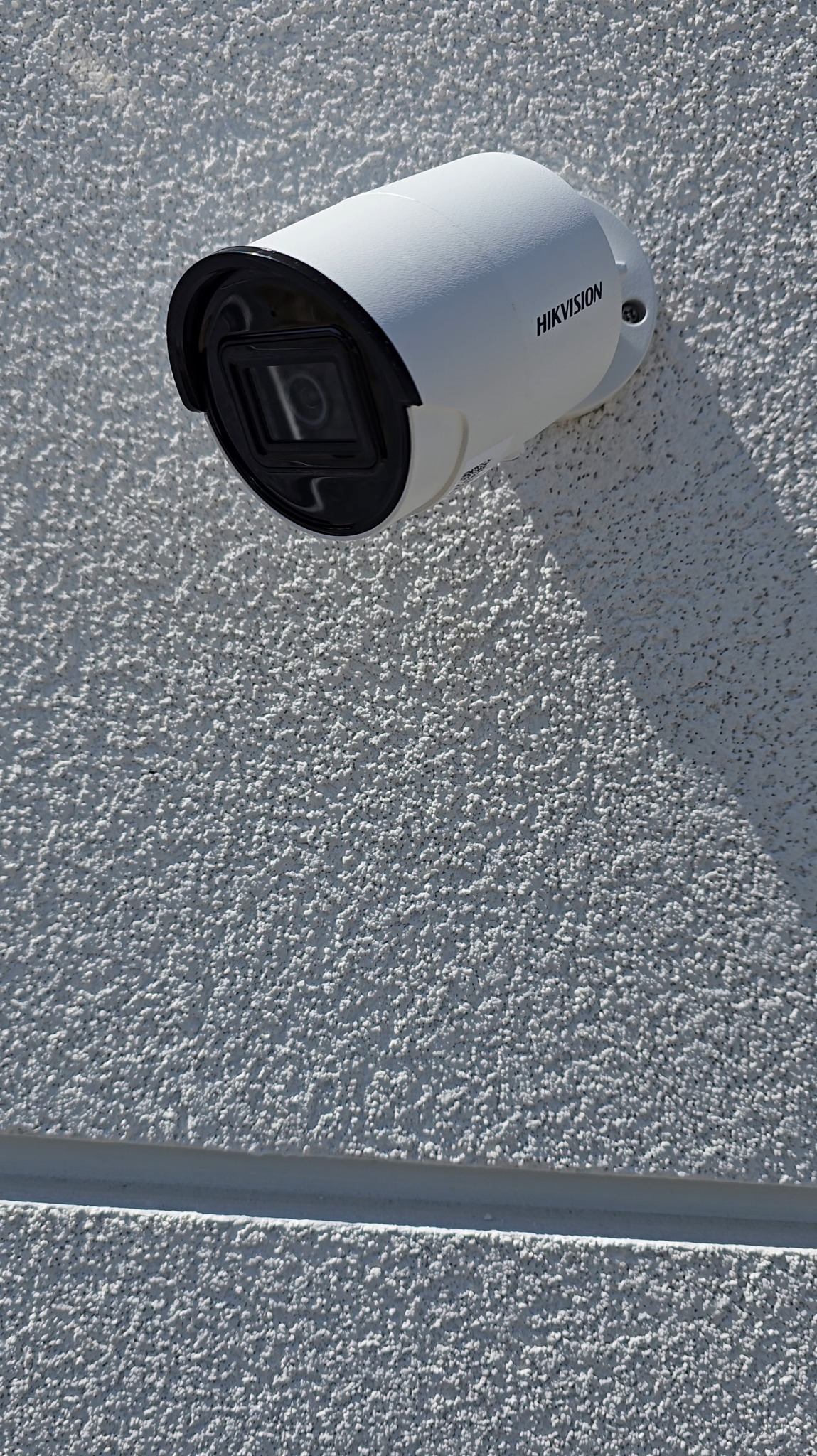 Biała kamera monitoringu Hikvision zamontowana na szarej, teksturowanej ścianie budynku. Widoczny cień kamery. Kadr pionowy, zbliżenie na kamerę.