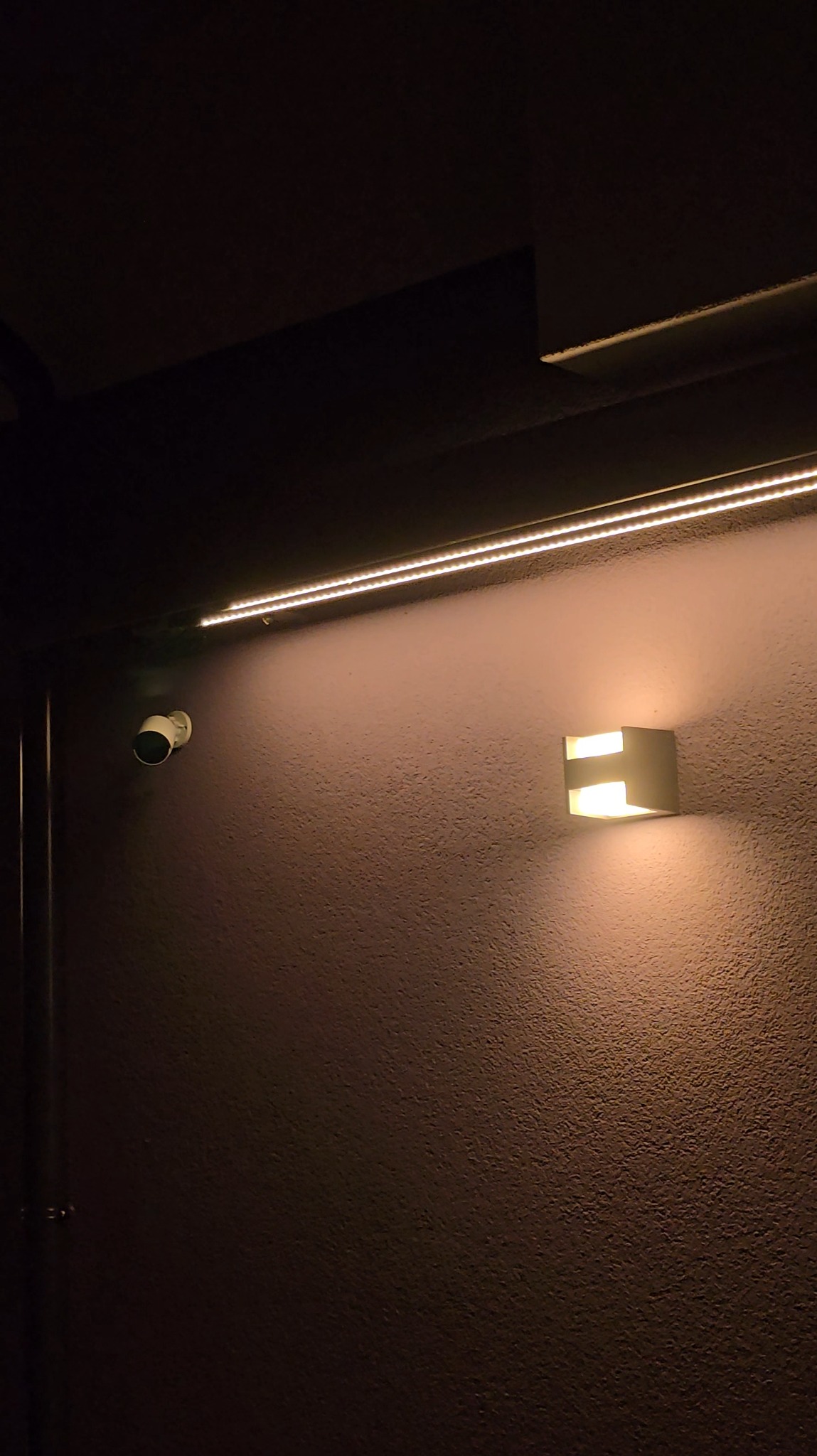 Kamera monitoringu, listwa LED i nowoczesny kinkiet na teksturowanej ścianie w ciemnym pomieszczeniu. Oświetlenie akcentujące.