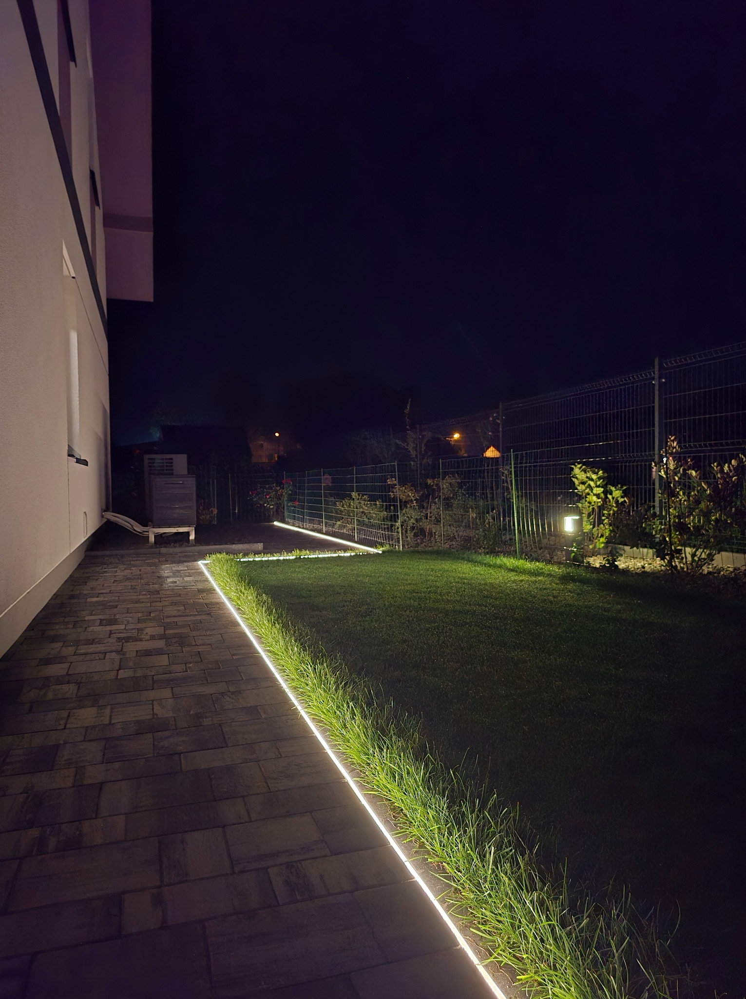 Oświetlenie LED wzdłuż ścieżki i trawnika przy domu, nocne ujęcie. Subtelne, liniowe światło podkreśla architekturę ogrodu.
