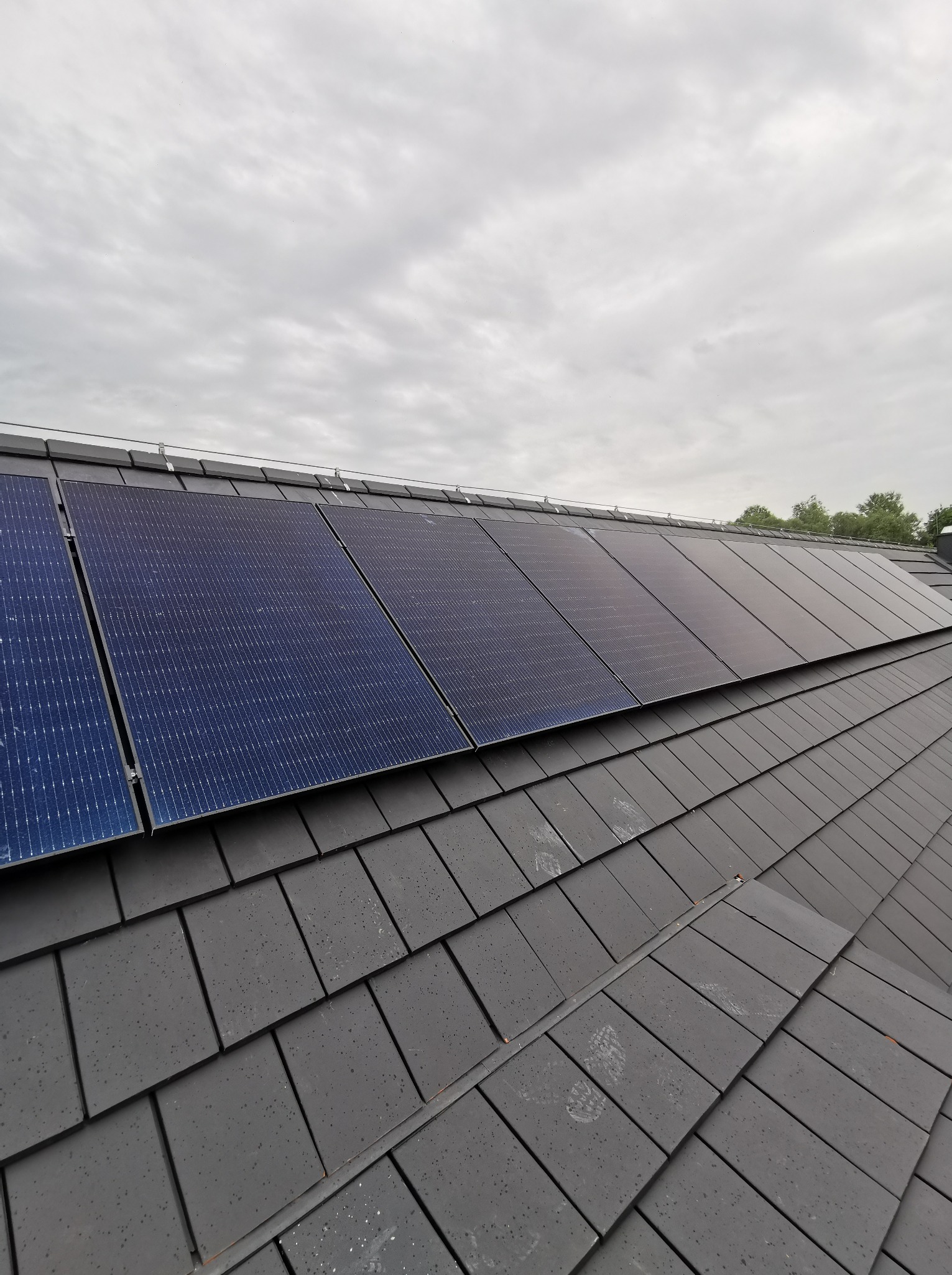 Panele słoneczne zamontowane na dachu pokrytym szarą dachówką, widok z dołu pod kątem, pochmurne niebo w tle. Widoczne ślady na dachówce.