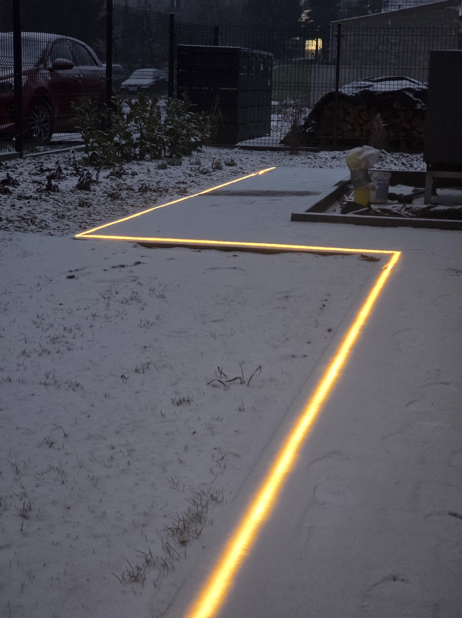 Świecące linie LED w śniegu wyznaczają ścieżkę w ogrodzie. Widoczne krzewy, ogrodzenie i zaparkowany samochód w tle. Nietypowe oświetlenie ogrodu zimą.