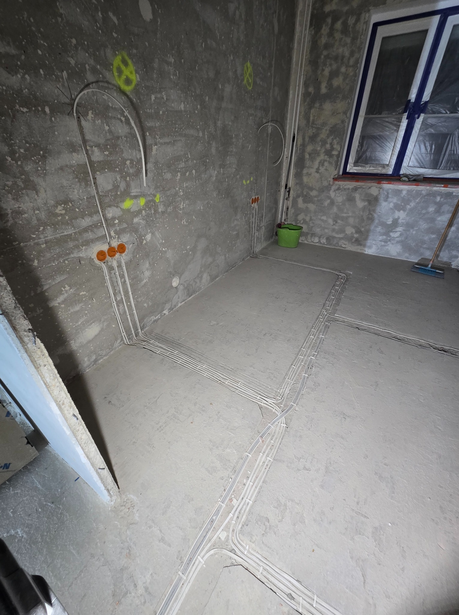Surowy pokój z widoczną instalacją elektryczną w podłodze i ścianach, przygotowany do dalszych prac wykończeniowych, okno zabezpieczone folią.