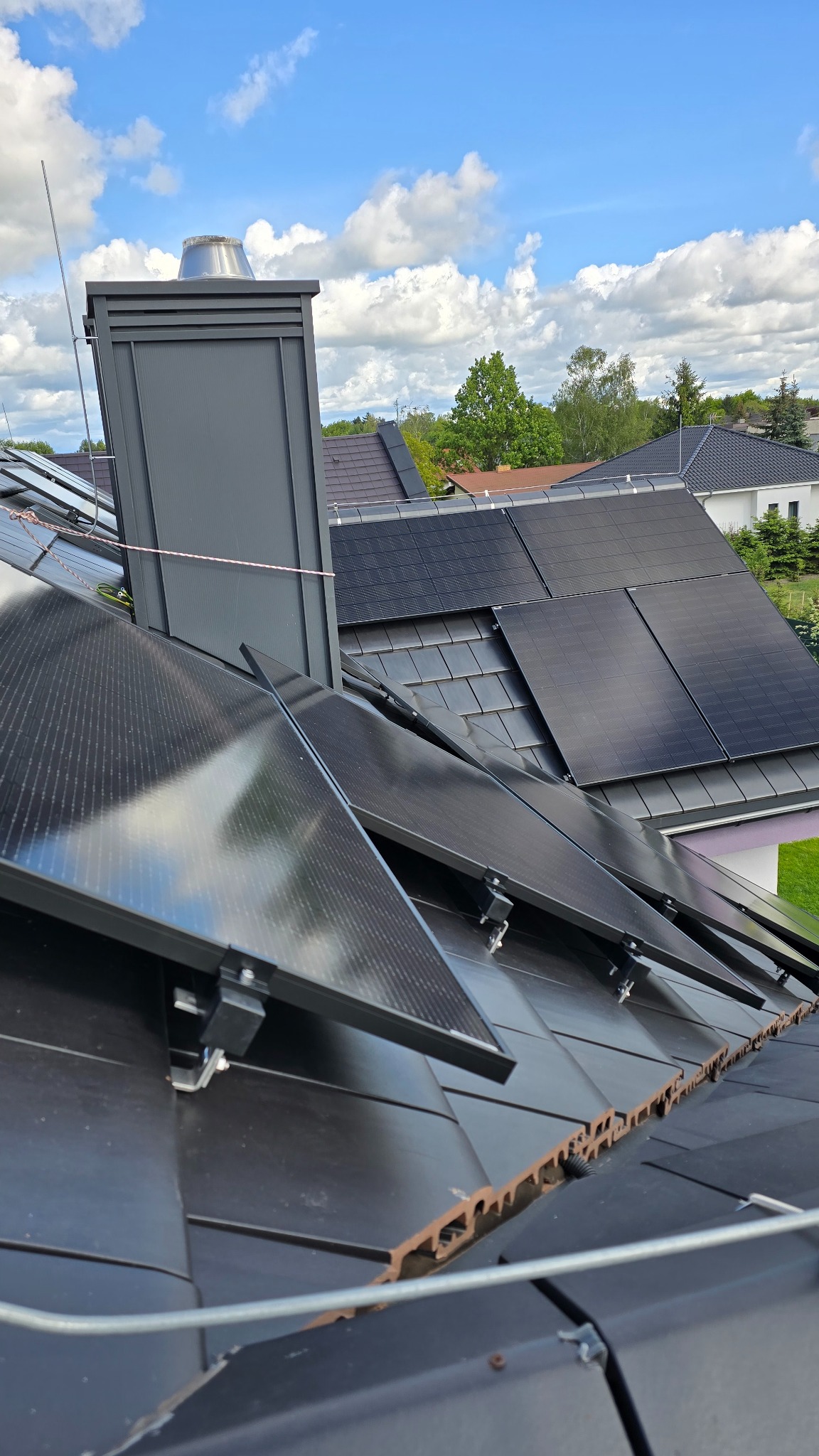 Instalacja paneli fotowoltaicznych na dachu z czarnej dachówki. Widoczny komin i instalacja odgromowa. Błękitne niebo z chmurami w tle.