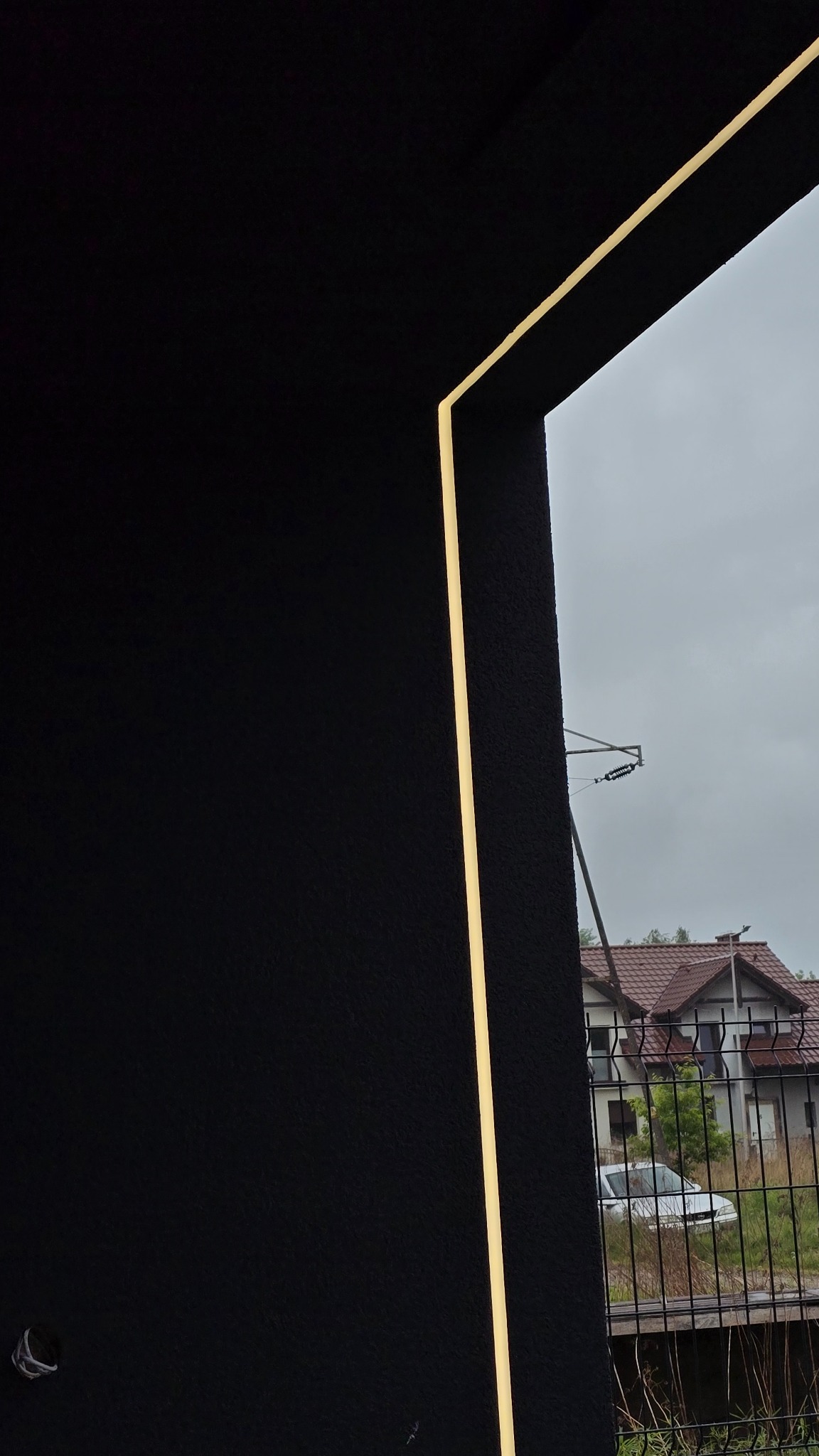 Elegancka, pionowa oprawa oświetleniowa na czarnej elewacji budynku, widok z dołu na szarym tle nieba i fragment domu za ogrodzeniem.