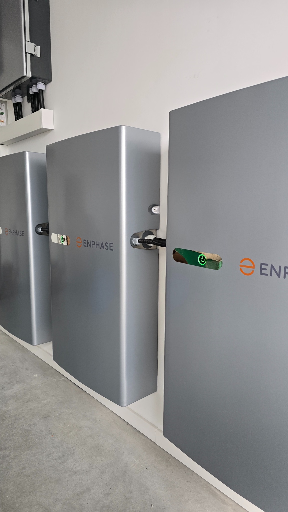 Szereg szarych modułów Enphase z przewodami, zamontowanych na białej ścianie, z widocznym logo i wskaźnikami LED, sugerujących system zasilania lub magazynowania energii.