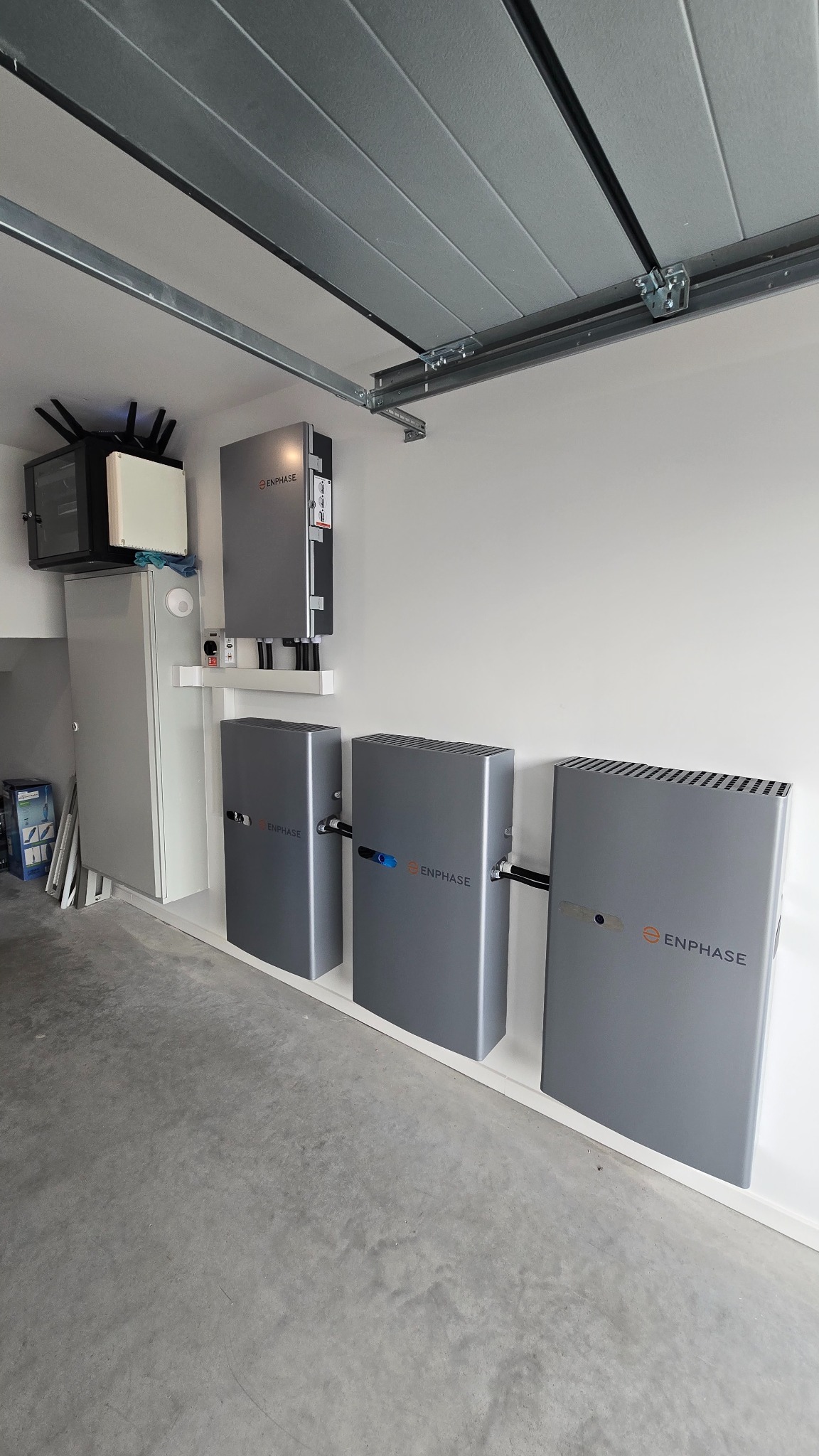 Szara instalacja Enphase w garażu: trzy baterie podłączone przewodami, moduł sterujący na ścianie, szafka z routerem. Nowoczesny system zasilania.