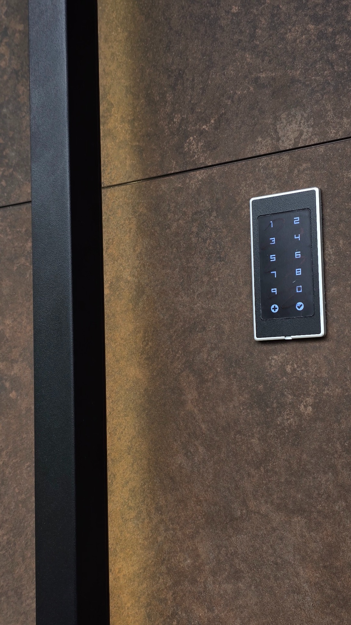 Elegancki panel dotykowy domofonu z numeracją na tle ściany o fakturze rdzy i fragmentu czarnej ramy, nowoczesne rozwiązanie kontroli dostępu.