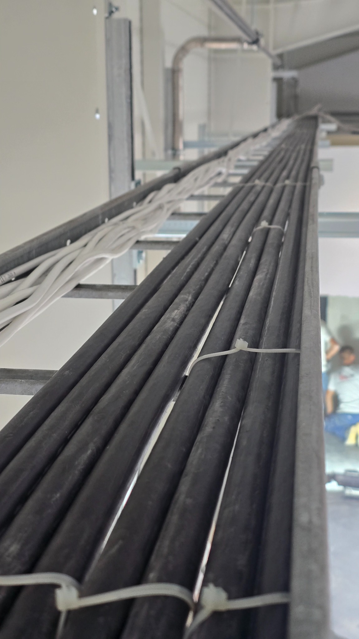 Ułożone równolegle kable w szarych peszlach, przymocowane do metalowej konstrukcji w jasnym wnętrzu. Widok z dołu, perspektywa zbieżna, białe kable w tle.