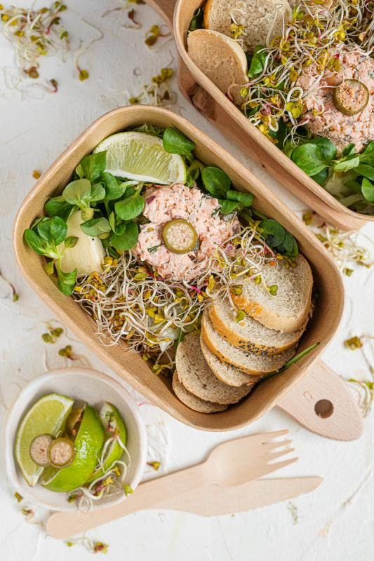 Lunchbox z sałatką z łososia, kiełkami, sałatą i limonką. Obok drugi lunchbox i miseczka z limonką. Drewniane sztućce. Zdrowy catering dietetyczny w ekologicznym opakowaniu.
