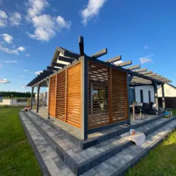Drewniana pergola z lamelami, szara konstrukcja, schody z kostki brukowej, widok na ogród i błękitne niebo z chmurami. Estetyczne wykończenie.