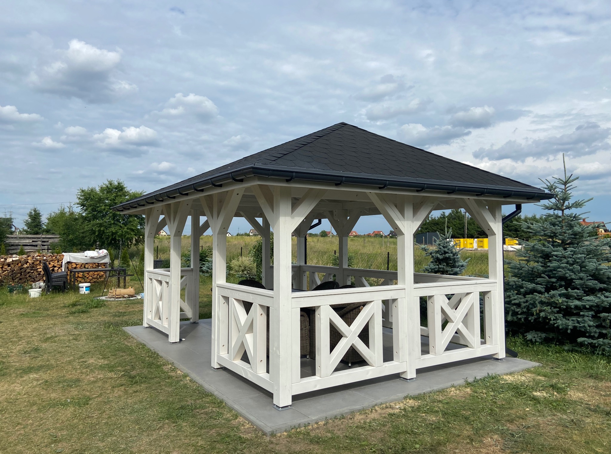Biała, drewniana altana z czarnym dachem na betonowej podstawie, otoczona zielenią i drzewami, pod błękitnym niebem z chmurami. Widok z przodu, w tle ułożone drewno.