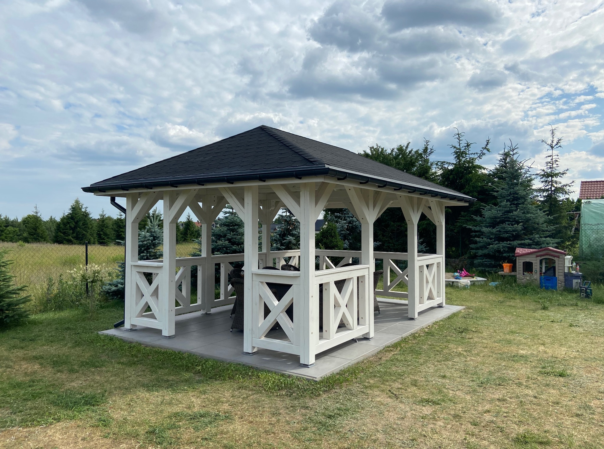 Biała altana ogrodowa z czarnym dachem na szarym podłożu. W tle zielona trawa, drzewa iglaste i pochmurne niebo. Widoczne elementy wyposażenia wnętrza altany.