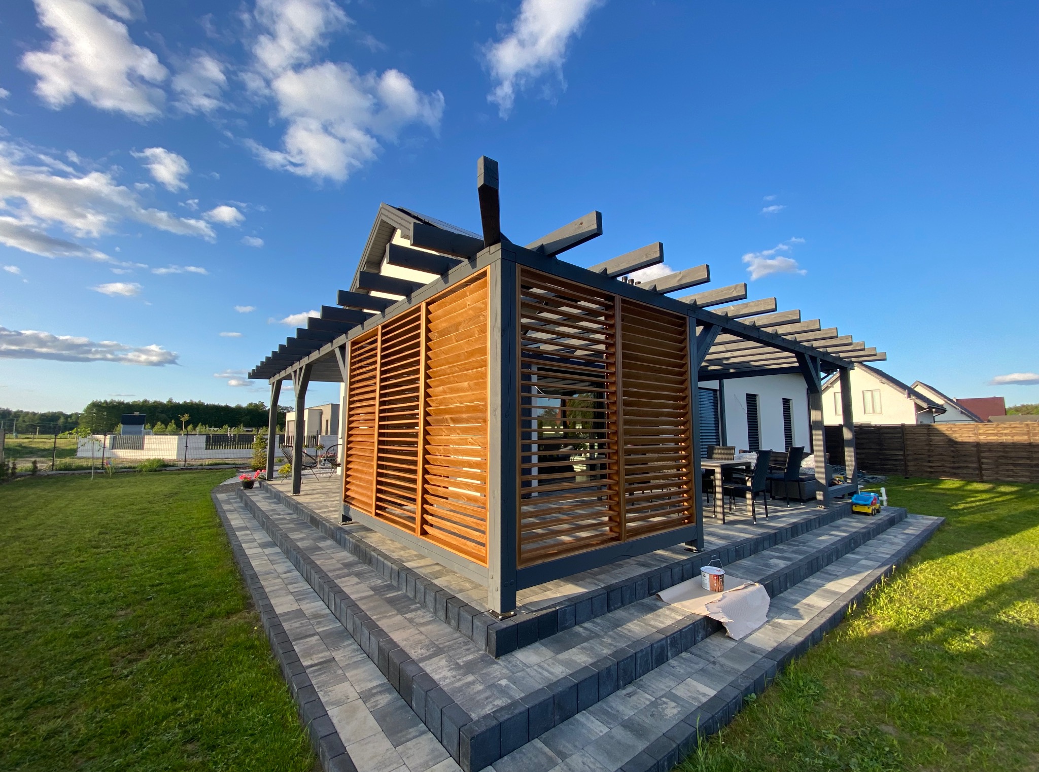 Drewniana pergola z lamelami, szara konstrukcja, schody z kostki brukowej, widok na ogród i błękitne niebo z chmurami. Estetyczne wykończenie.