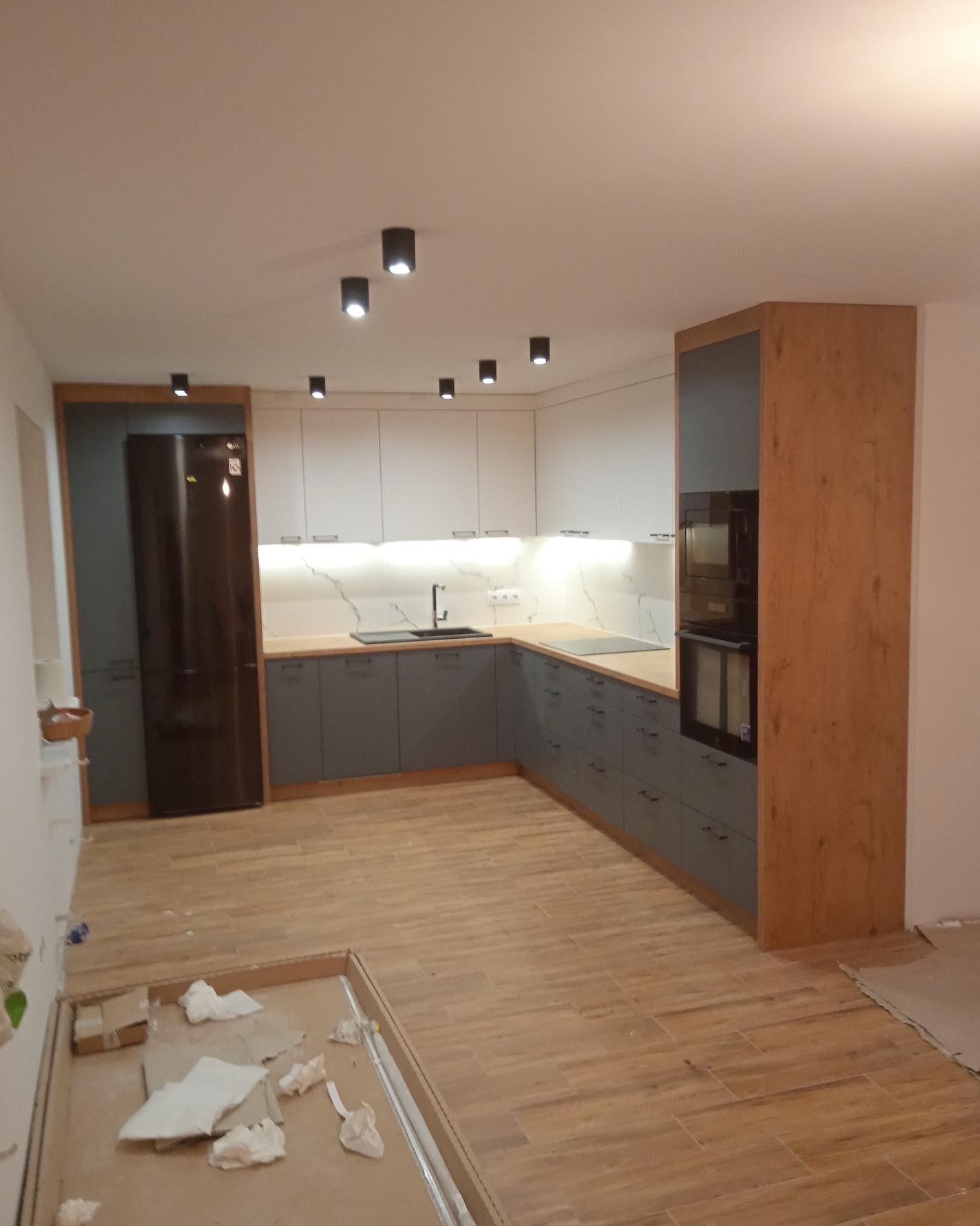 Nowoczesna kuchnia z szarymi szafkami dolnymi i białymi górnymi, drewnianym wykończeniem i oświetleniem punktowym, widok na podłogę i elementy wykończeniowe.