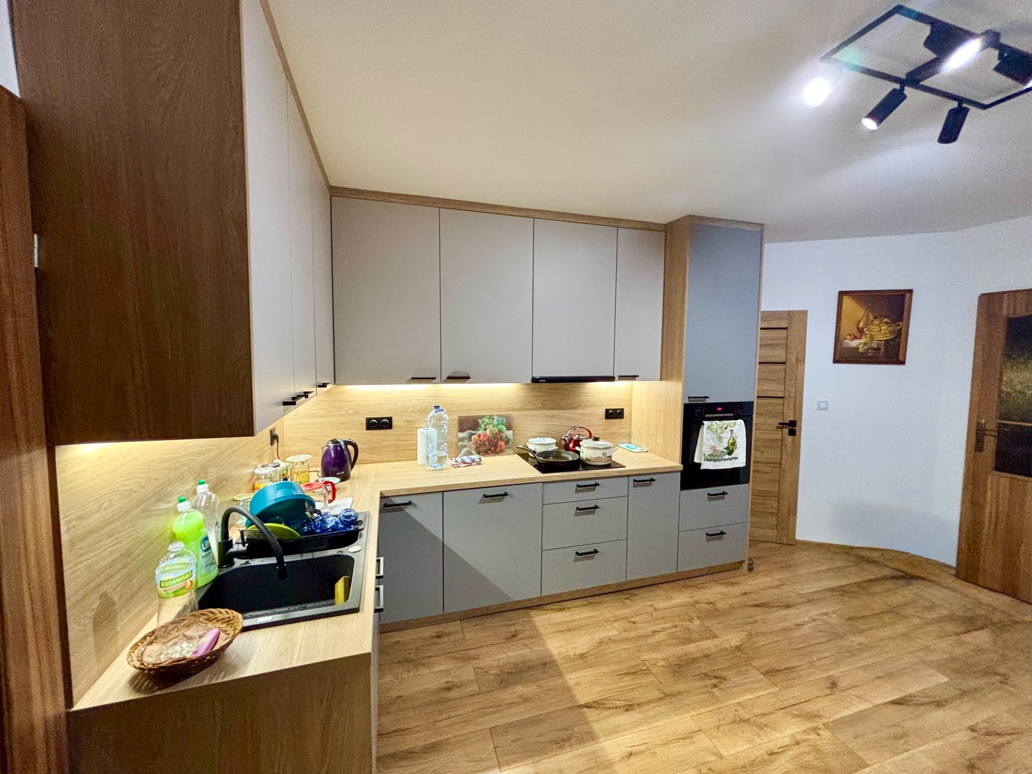 Jasna kuchnia z szarymi frontami i drewnianymi blatami, oświetlona listwami LED pod szafkami wiszącymi. Widoczny zlew z naczyniami i drewniana podłoga.