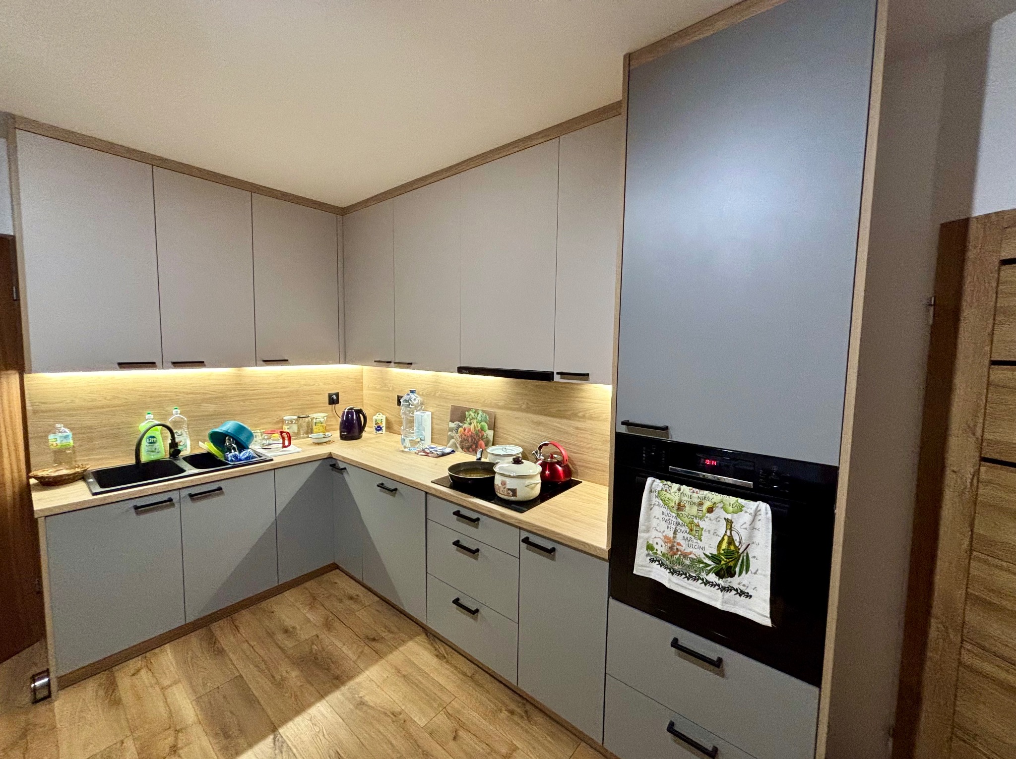 Nowoczesna kuchnia z szarymi frontami, drewnianym blatem i czarnymi uchwytami. Na blacie naczynia, czajnik i płyta grzewcza. W tle widoczne oświetlenie podszafkowe.