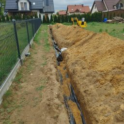 USŁUGI OGÓLNOBUDOWLANE SEBASTIAN KOCIĘCKI - Wykopany rów z czarną folią w ziemi, obok ogrodzenie panelowe, w tle domy i minikoparka. Widok na instalację w trakcie prac ziemnych.