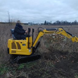 USŁUGI OGÓLNOBUDOWLANE SEBASTIAN KOCIĘCKI - Operator na żółtej minikoparce GG800 w trakcie prac ziemnych na zewnątrz, z widocznymi śladami wykopu. Tło: pole i zabudowania w oddali pod pochmurnym niebem.