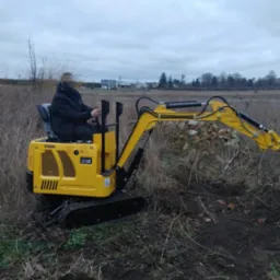Operator na żółtej minikoparce GG800 w trakcie prac ziemnych na zewnątrz, z widocznymi śladami wykopu. Tło: pole i zabudowania w oddali pod pochmurnym niebem.