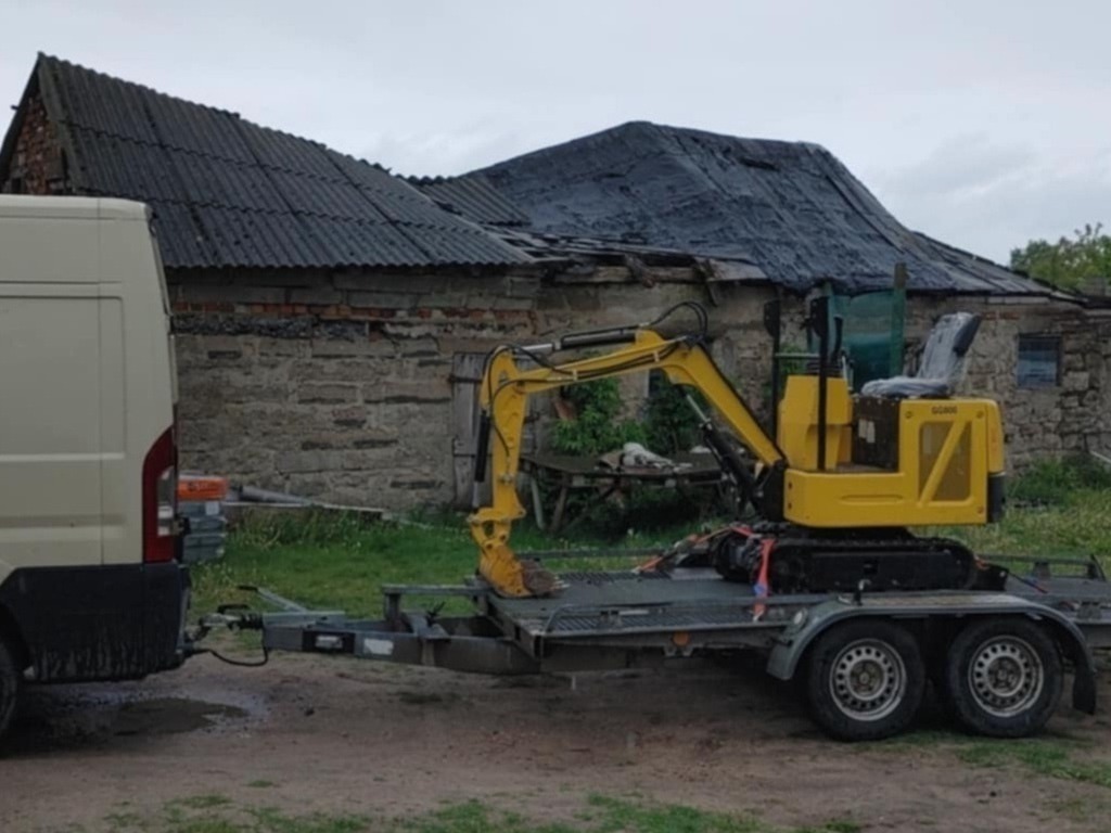 Żółta mini koparka SCM86 na lawecie, podpięta do busa. W tle stary, kamienny budynek z dziurawym dachem. Sprzęt gotowy do transportu na miejsce wykopu.