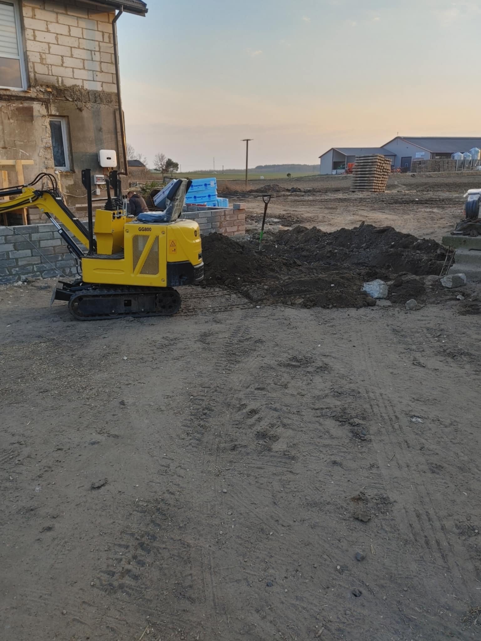 Żółta mini koparka GG800 na placu budowy, obok wykopu z ziemią i wystającymi prętami zbrojeniowymi. W tle budynek w trakcie budowy i zabudowania gospodarcze.