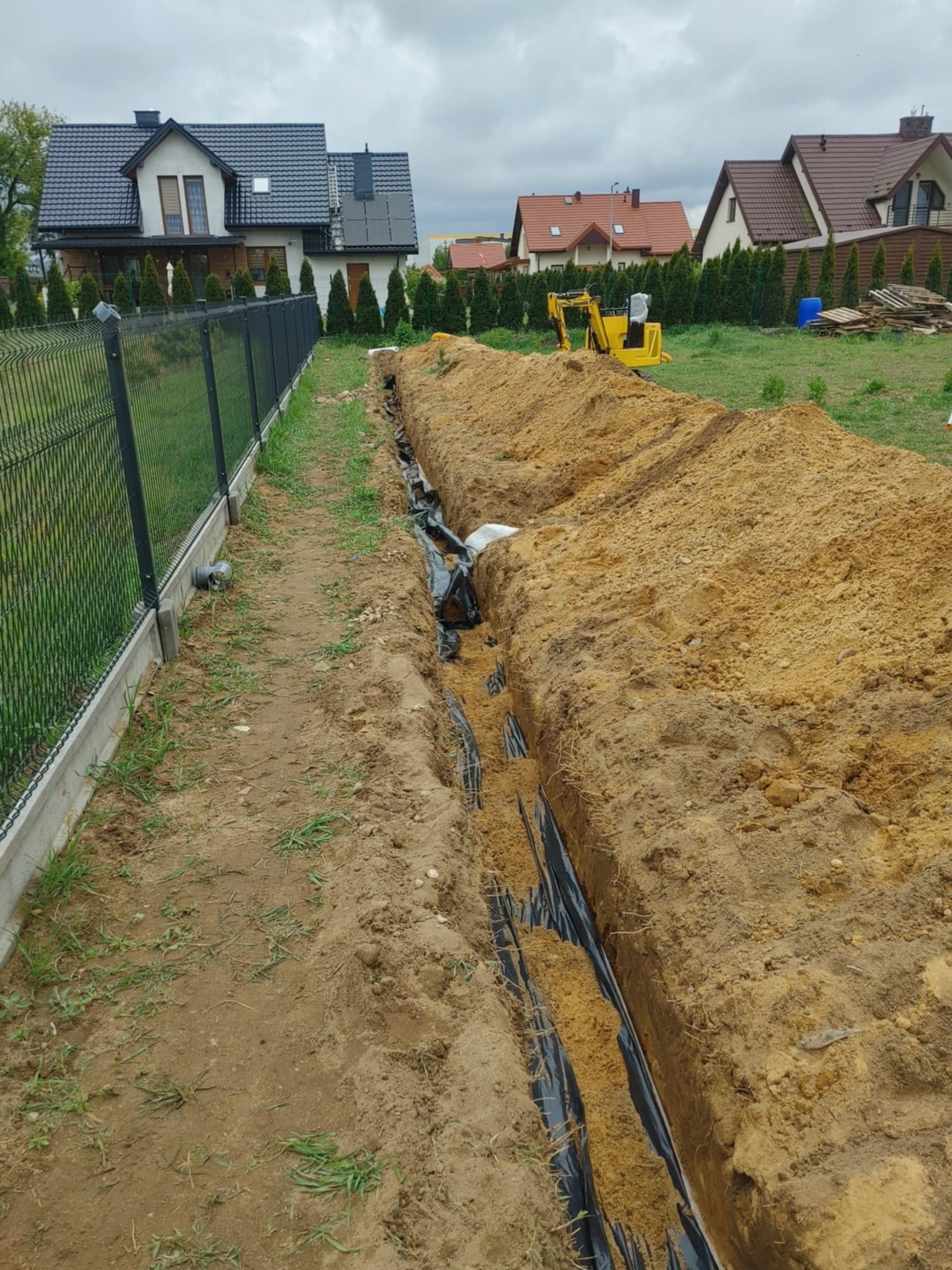 Wykopany rów z czarną folią w ziemi, obok ogrodzenie panelowe, w tle domy i minikoparka. Widok na instalację w trakcie prac ziemnych.
