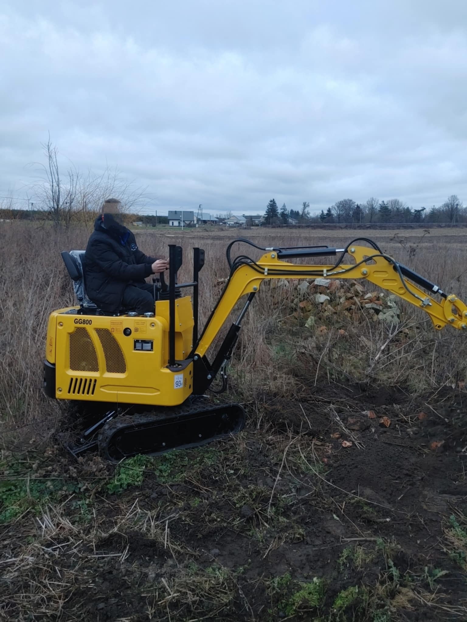 Operator na żółtej minikoparce GG800 w trakcie prac ziemnych na zewnątrz, z widocznymi śladami wykopu. Tło: pole i zabudowania w oddali pod pochmurnym niebem.