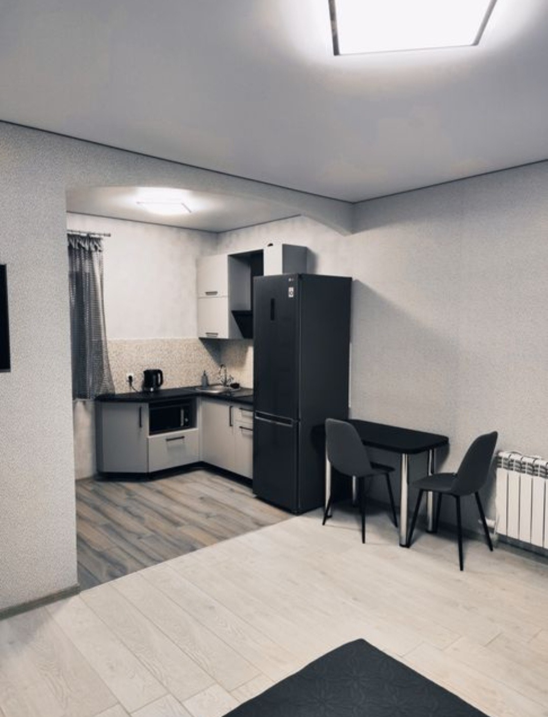 Wnętrze pokoju z aneksem kuchennym w odcieniach szarości i bieli. Widoczne meble kuchenne, stół z krzesłami, lodówka i grzejnik. Minimalistyczny design.