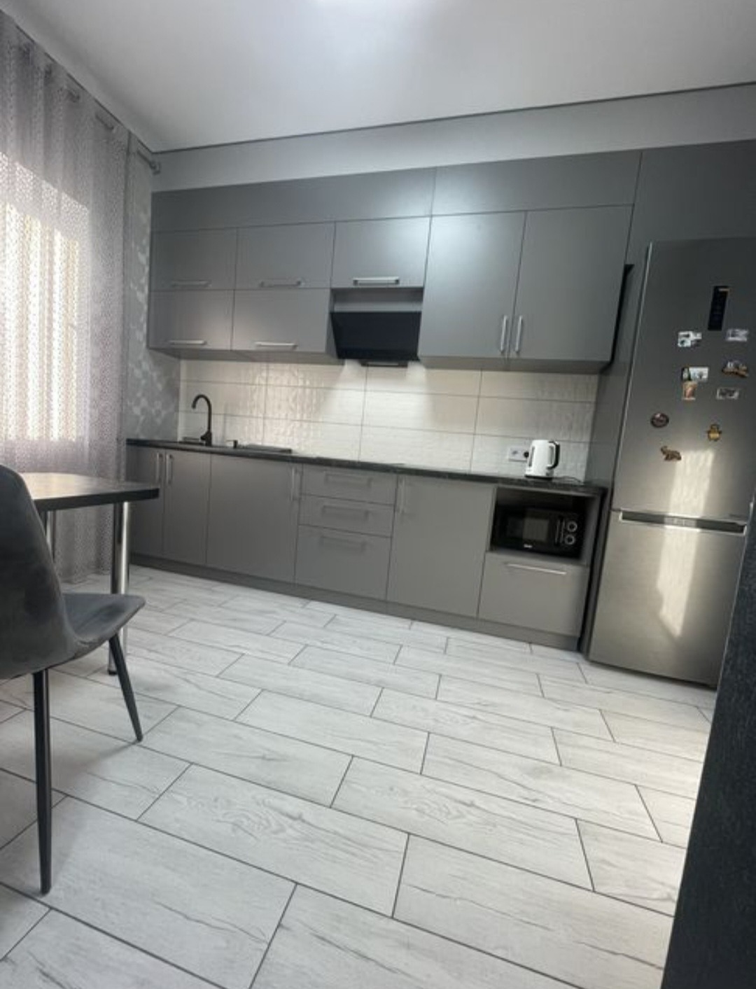 Nowoczesna, szara kuchnia z drewnopodobną podłogą i stołem z krzesłami. Minimalistyczny design, czyste linie, jasne wnętrze z oknem z lewej strony.