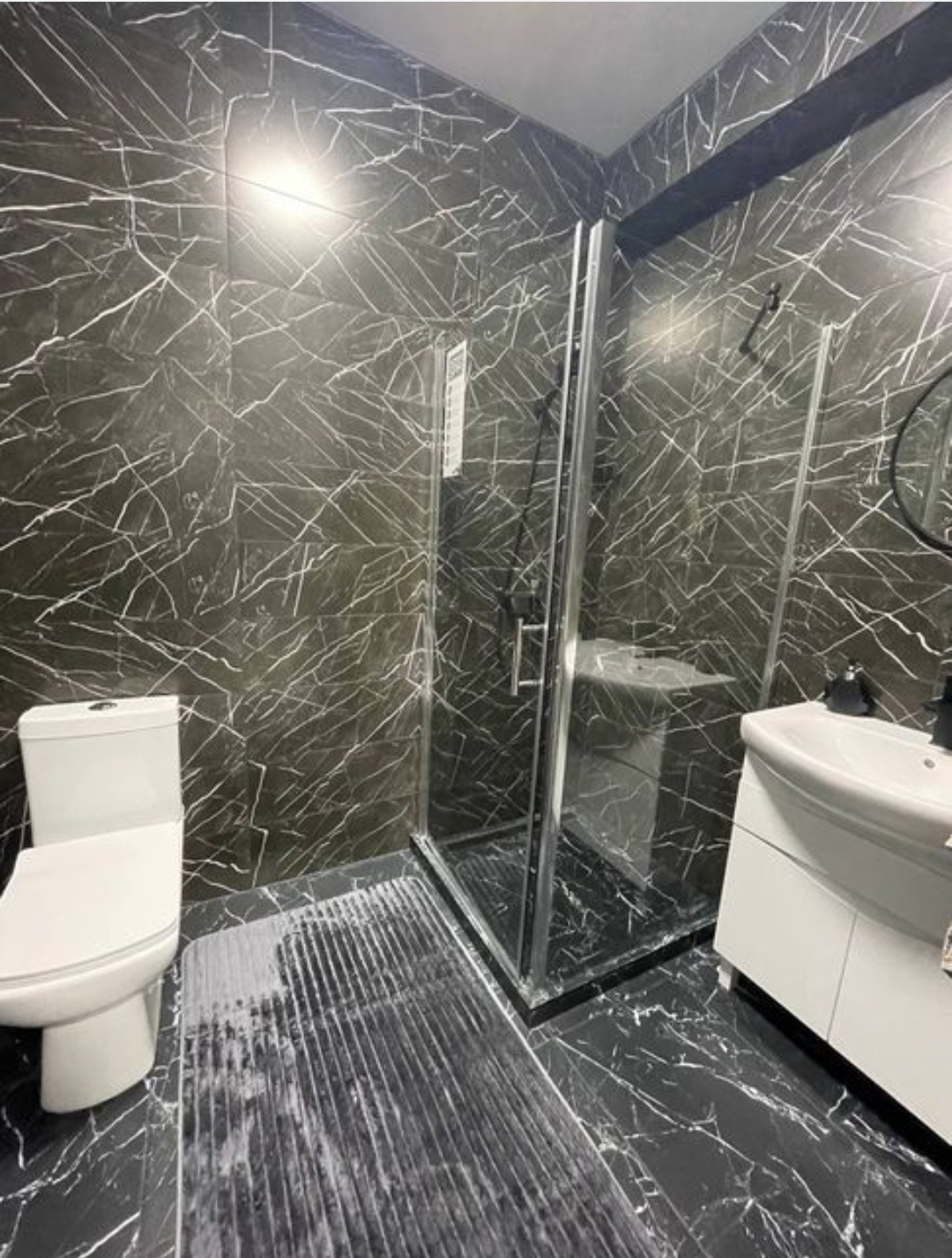 Wykończona łazienka z kabiną prysznicową, toaletą i umywalką na szafce. Czarno-białe płytki imitujące marmur na ścianach i podłodze. Nowoczesny design.