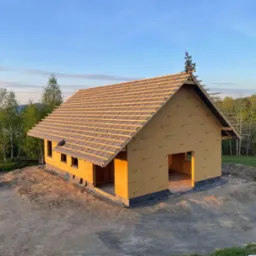Budowa sauny: szkielet konstrukcji z drewnianym dachem w trakcie montażu, widok z zewnątrz na tle zieleni i błękitnego nieba o zmierzchu. Etap budowy, bez wykończenia.