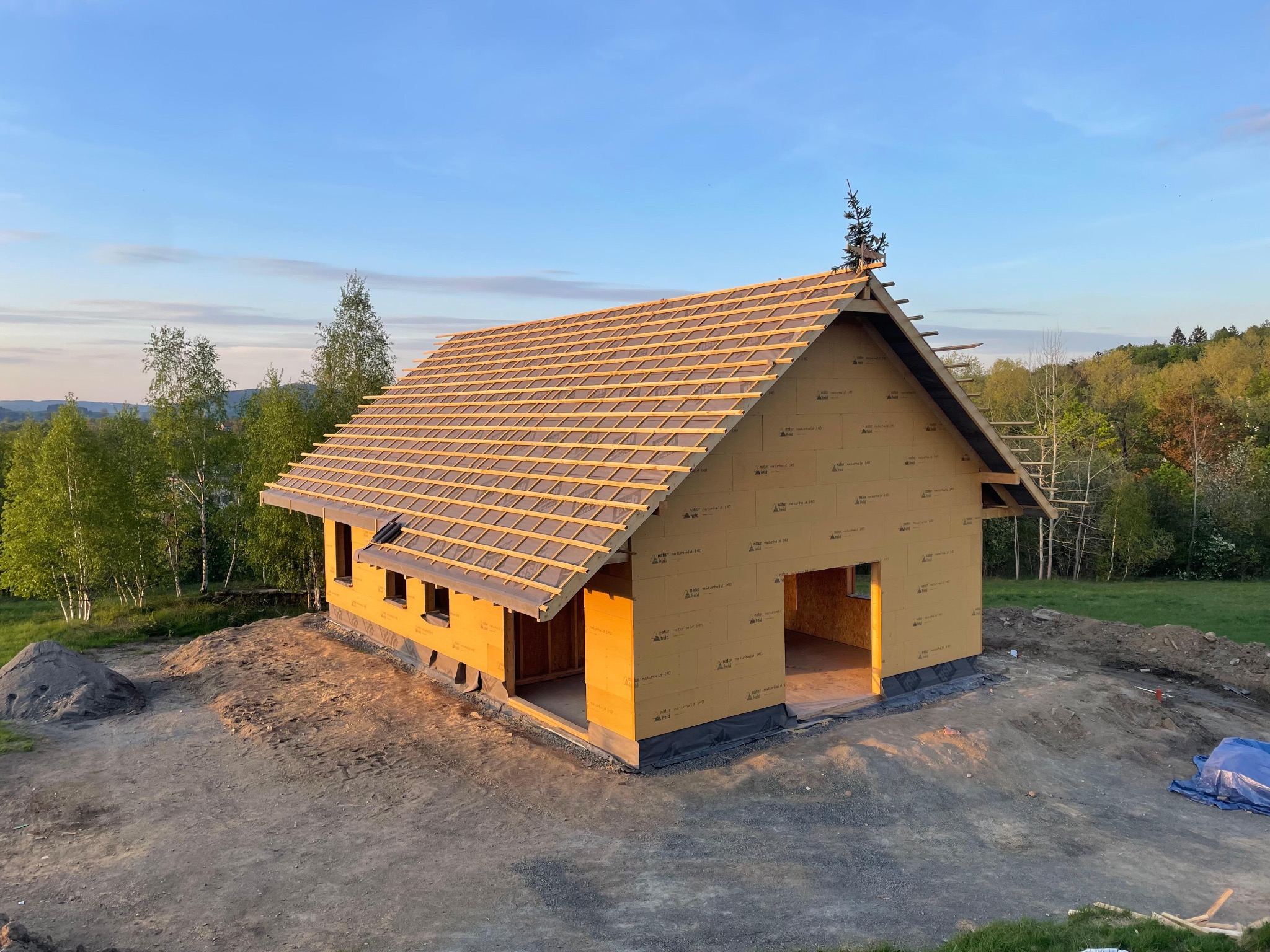 Budowa sauny: szkielet konstrukcji z drewnianym dachem w trakcie montażu, widok z zewnątrz na tle zieleni i błękitnego nieba o zmierzchu. Etap budowy, bez wykończenia.