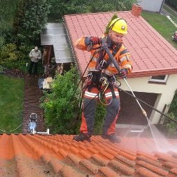 TODO BARTOSZ SZENDZIKOWSKI - Pracownik w kasku i uprzęży myje dachówkę myjką ciśnieniową w Łodzi. Widok z góry ukazuje czyszczony fragment dachu i otoczenie budynku.
