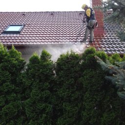 TODO BARTOSZ SZENDZIKOWSKI - Pracownik w kasku i uprzęży czyści dach z czerwonej dachówki za pomocą myjki ciśnieniowej. W tle komin z cegły i zielone drzewa.