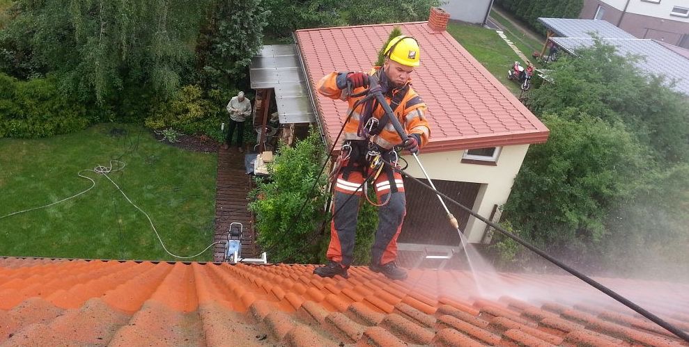 Pracownik w kasku i uprzęży myje dachówkę myjką ciśnieniową w Łodzi. Widok z góry ukazuje czyszczony fragment dachu i otoczenie budynku.