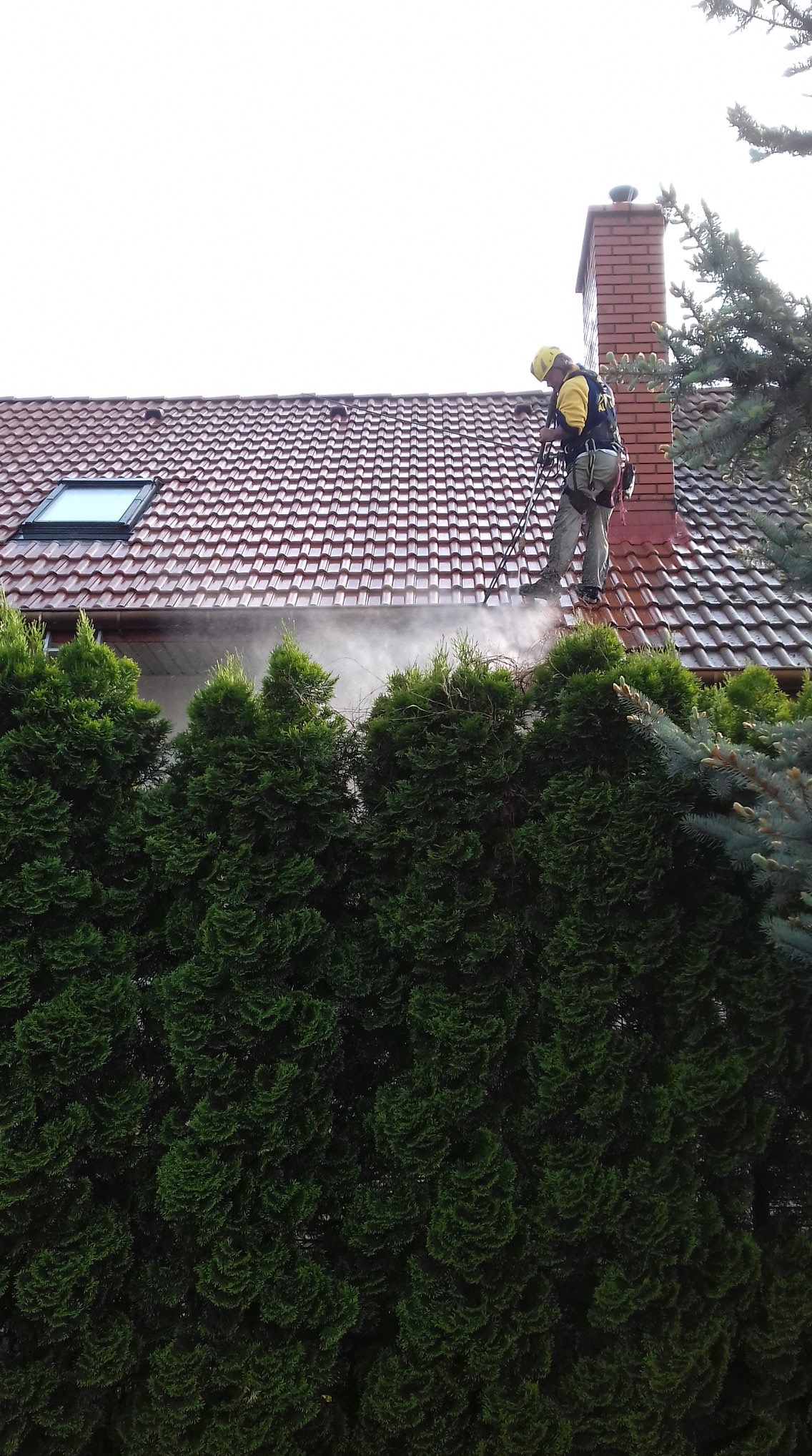 Pracownik w kasku i uprzęży czyści dach z czerwonej dachówki za pomocą myjki ciśnieniowej. W tle komin z cegły i zielone drzewa.