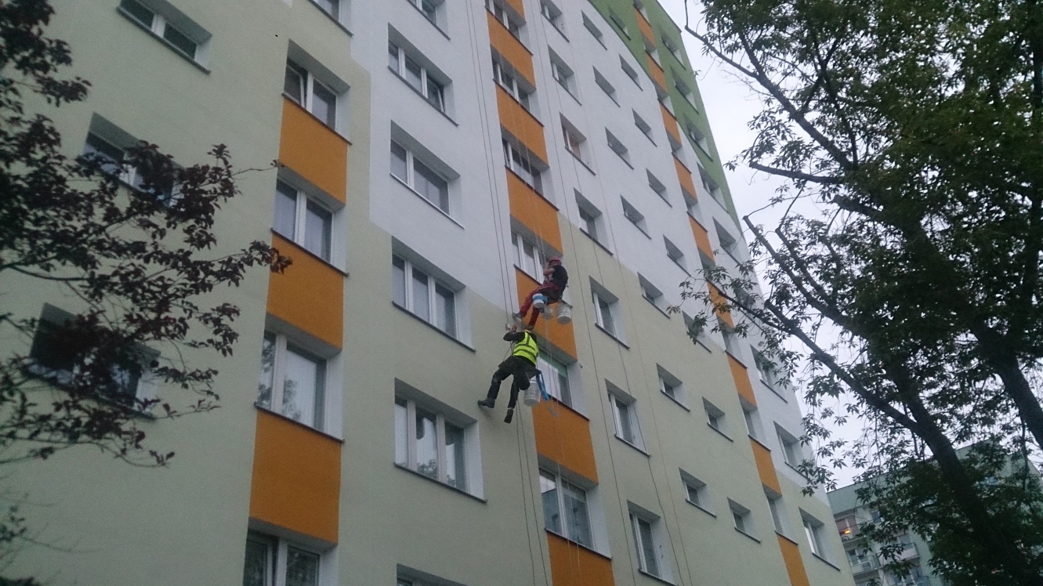 Praca na wysokości: dwuosobowy zespół alpinistyczny odświeża elewację bloku, używając lin i farb. Widoczne okna, fragmenty drzew i niebo w tle.